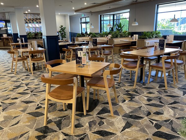 A spacious dining area with multiple wooden tables and chairs, a patterned floor, and large windows providing natural light. - Meadowbrook Hotel (Meadowbrook)
