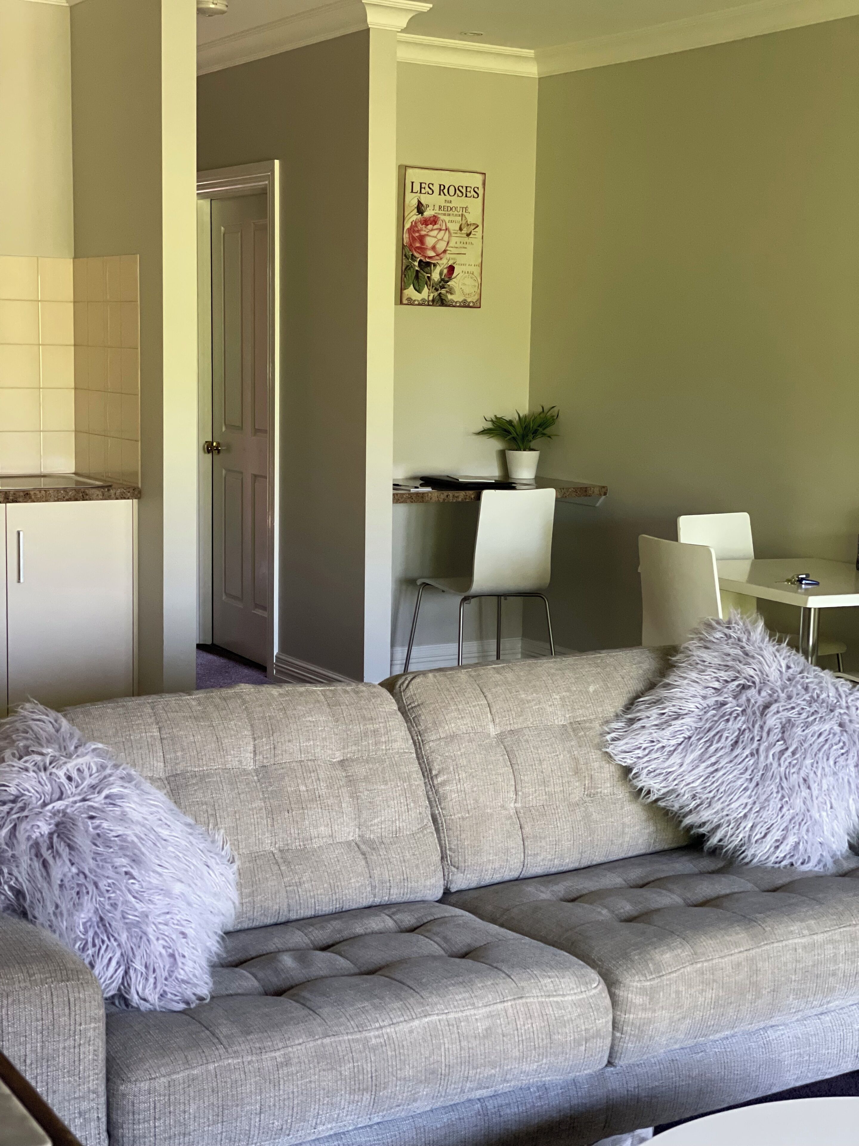 Een woonkamer met een grijze bank, twee zachte kussens, een eethoek met een witte tafel en stoelen, en een poster aan de muur.