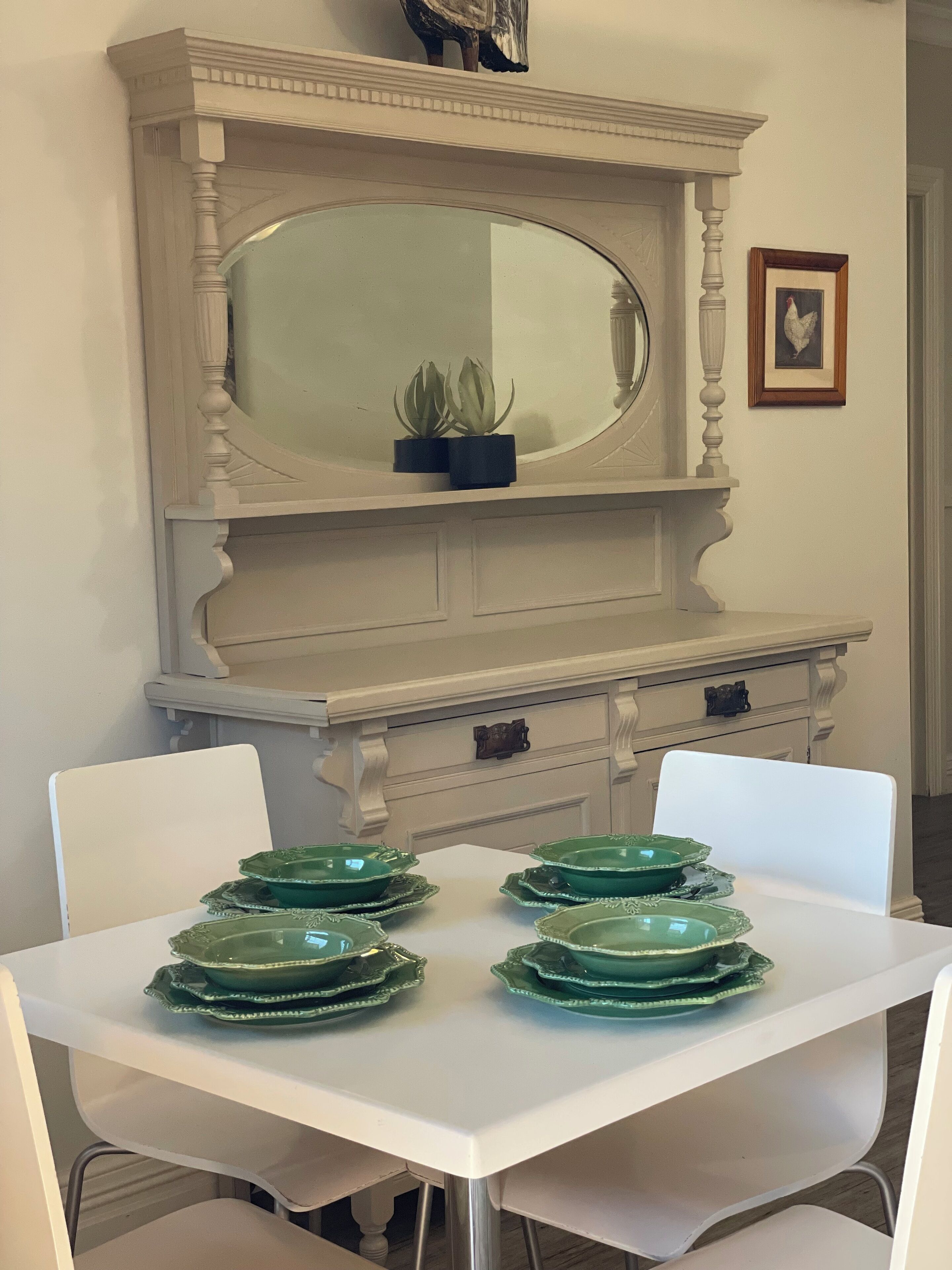 Un comedor con una mesa blanca dispuesta para cuatro, una silla blanca, un armario blanco con un espejo y un cuadro en la pared.