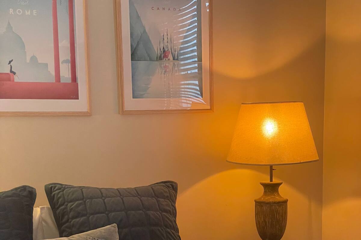 Una cama bien hecha con ropa oscura, dos almohadas decorativas, una lámpara y dos obras de arte enmarcadas en la pared.