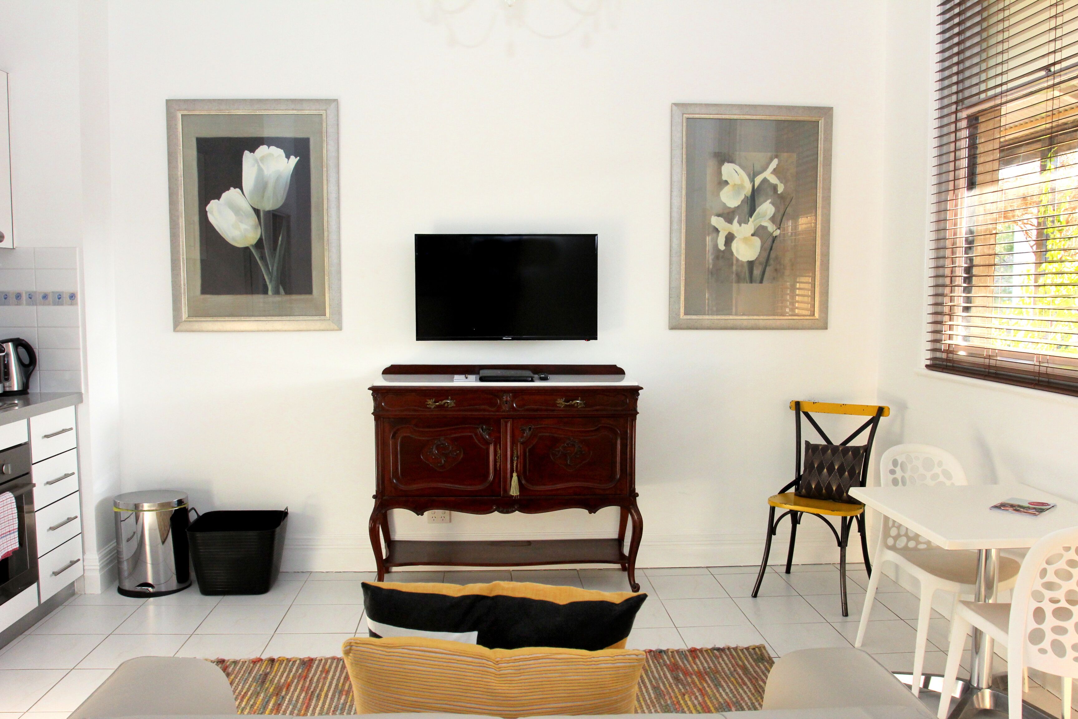 Ein Wohnzimmer mit Flachbildfernseher, einer Holzkommode, einem Esstisch mit Stühlen und zwei gerahmten floralen Bildern an der Wand.