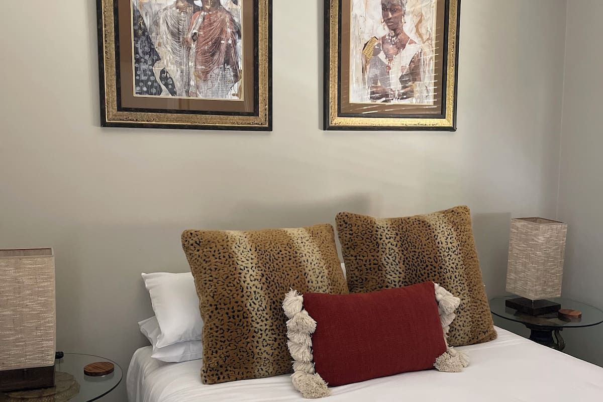 Un dormitorio con una cama, dos cuadros enmarcados, almohadas de estampado de leopardo, un cojín rojo y mesitas de noche con lámparas.