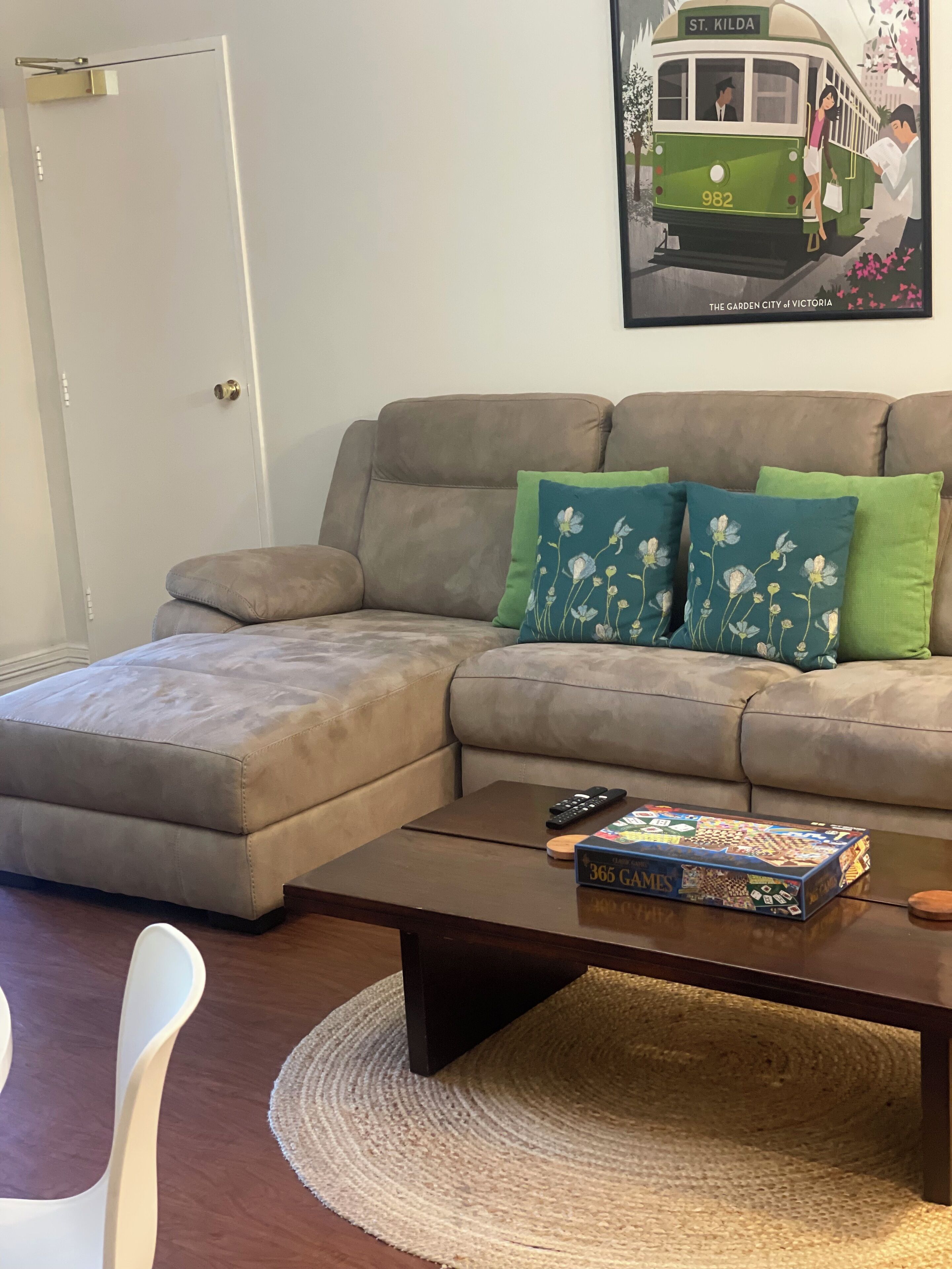 A living room with a grey sectional sofa, two green and one blue floral pillow, a wooden coffee table with a board game, and a framed picture of a green tram.