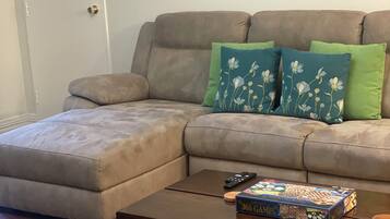 A living room with a grey sectional sofa, two green and one blue floral pillow, a wooden coffee table with a board game, and a framed picture of a green tram.
