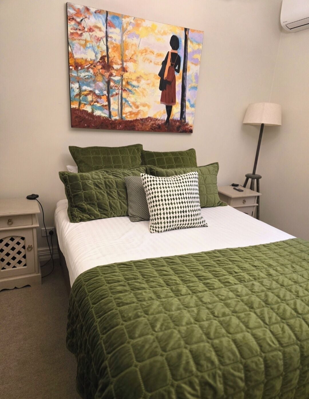 A neatly made bed with a green quilt and multiple pillows, a painting of a figure in a forest, a bedside lamp, and a nightstand with a remote control.