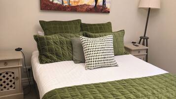 A neatly made bed with a green quilt and multiple pillows, a painting of a figure in a forest, a bedside lamp, and a nightstand with a remote control.