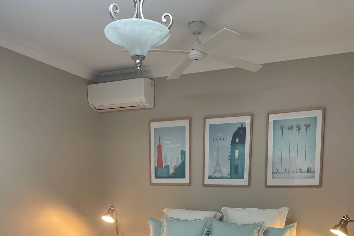 Un dormitorio con una cama, dos mesitas de noche, un ventilador de techo, un equipo de aire acondicionado y tres cuadros en la pared.