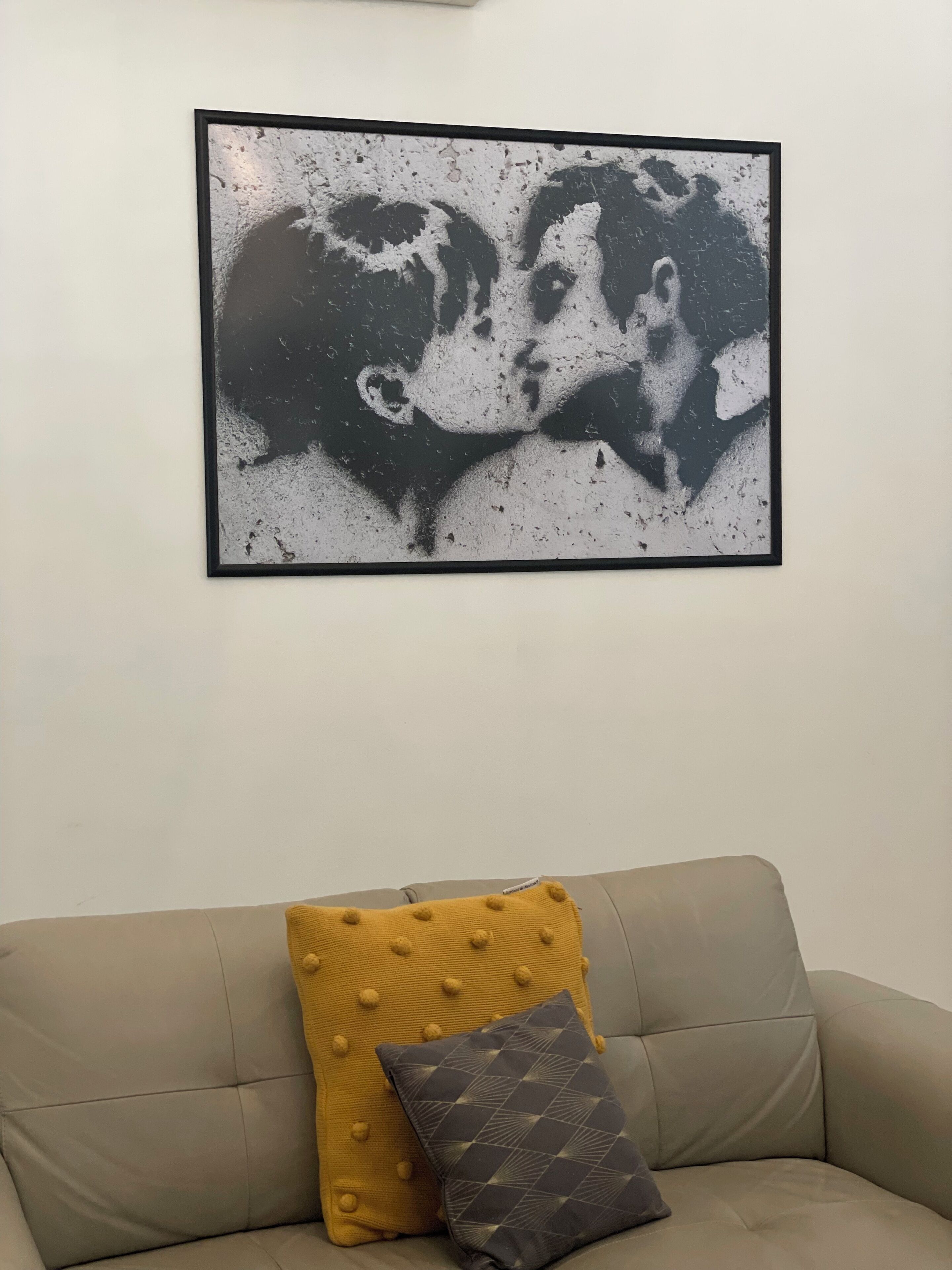 A framed artwork of a couple kissing on a white wall above a beige sofa with two cushions.