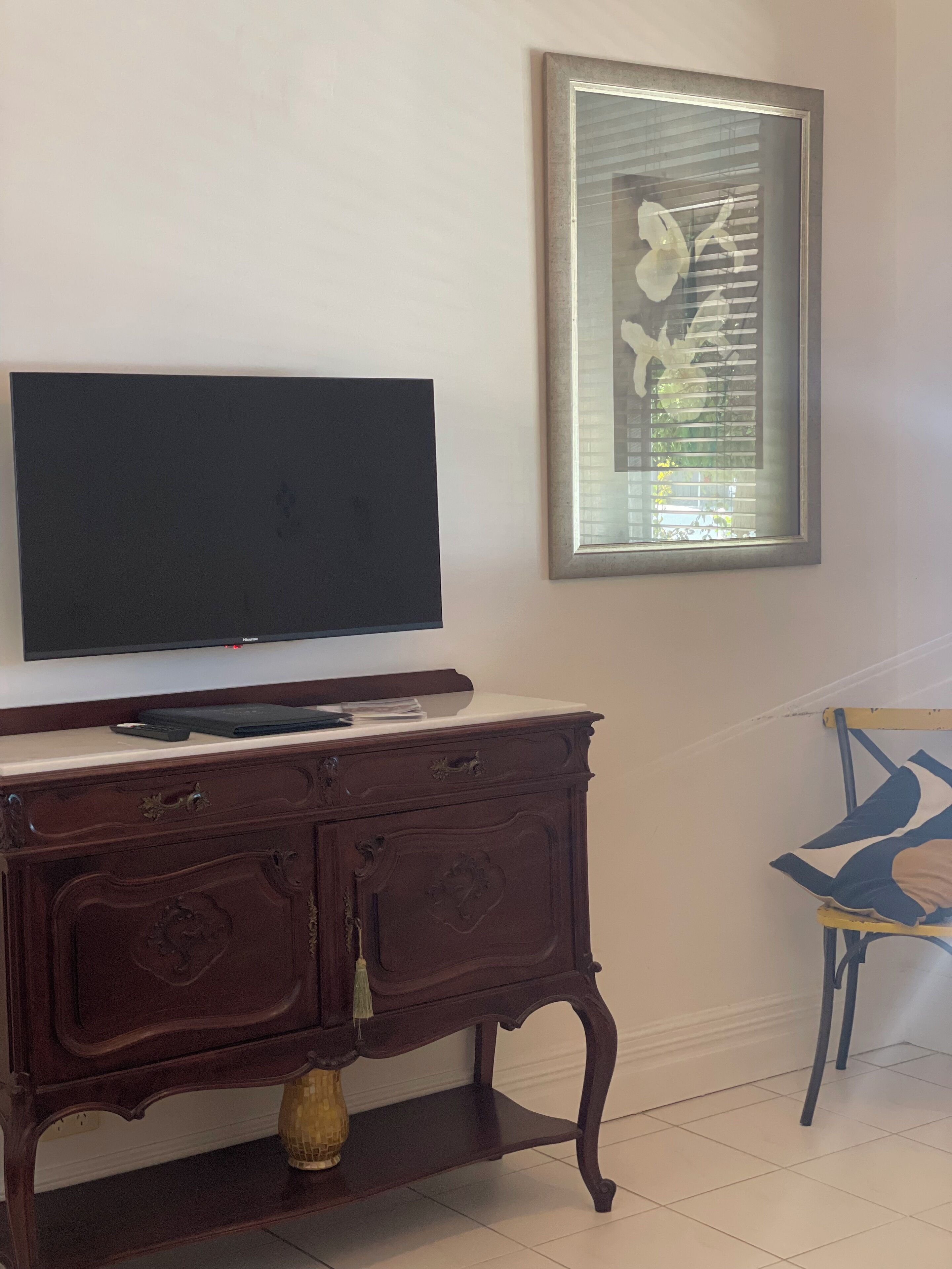 Una habitación con una televisión de pantalla plana montada en una pared blanca, un mueble de madera oscura con cajones, un pequeño florero decorativo y una obra de arte enmarcada sobre el mueble.
