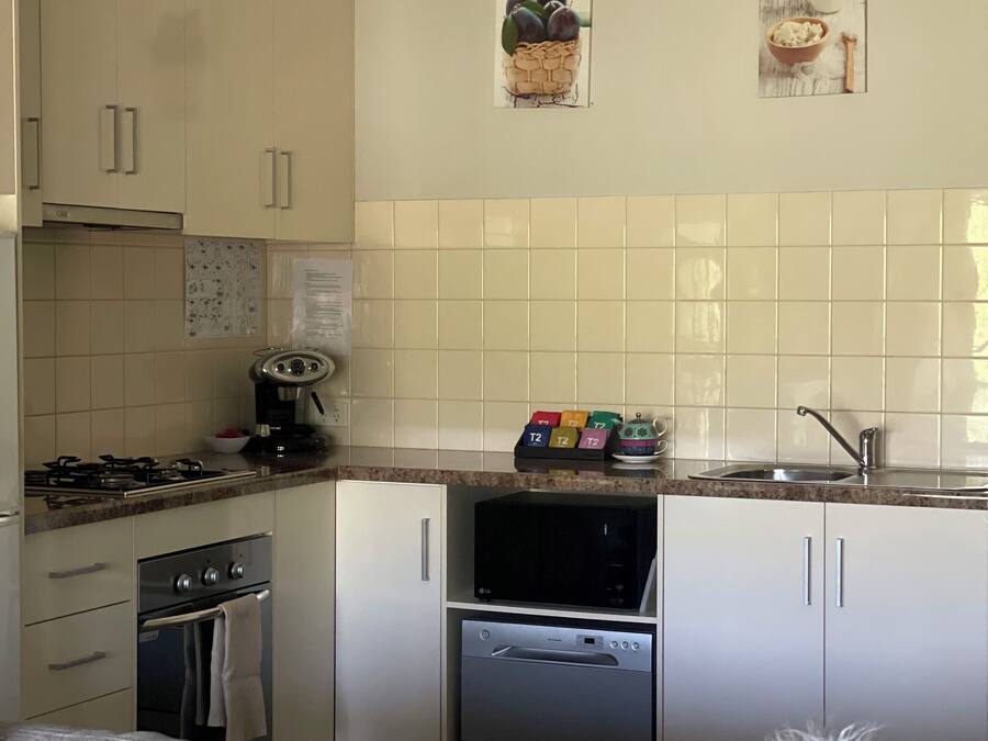 A compact kitchen with white cabinets, a gas stove, an oven, a microwave, and a dishwasher. There are two wall-mounted pictures and a set of colorful T2 boxes on the countertop.