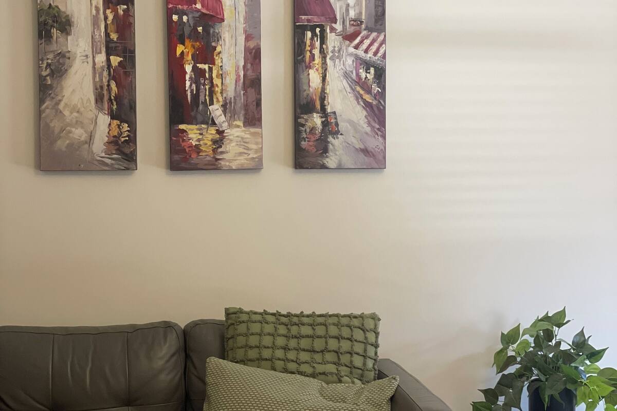 Un salón con un sofá de cuero marrón, dos cojines estampados y tres cuadros abstractos en la pared.