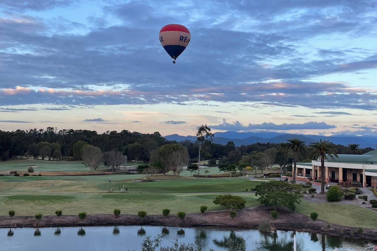 A hot air balloon over a golf course with a body of water in the foreground and a building in the background.