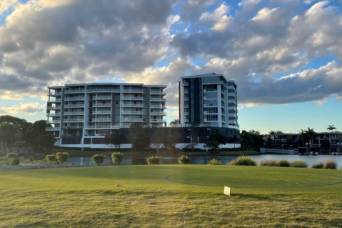 A modern multi-story building with balconies, a grassy area in front, and a body of water nearby.