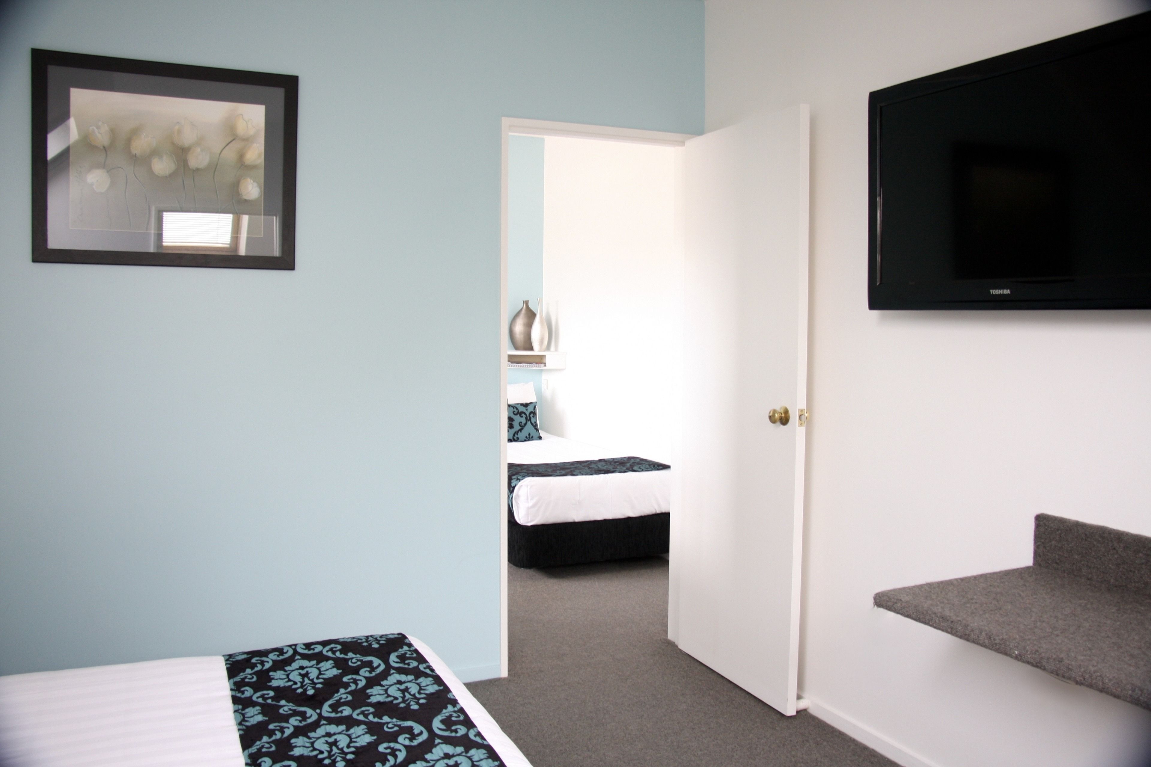 A hotel room with a bed, a TV, a framed artwork, and a door leading to another room.