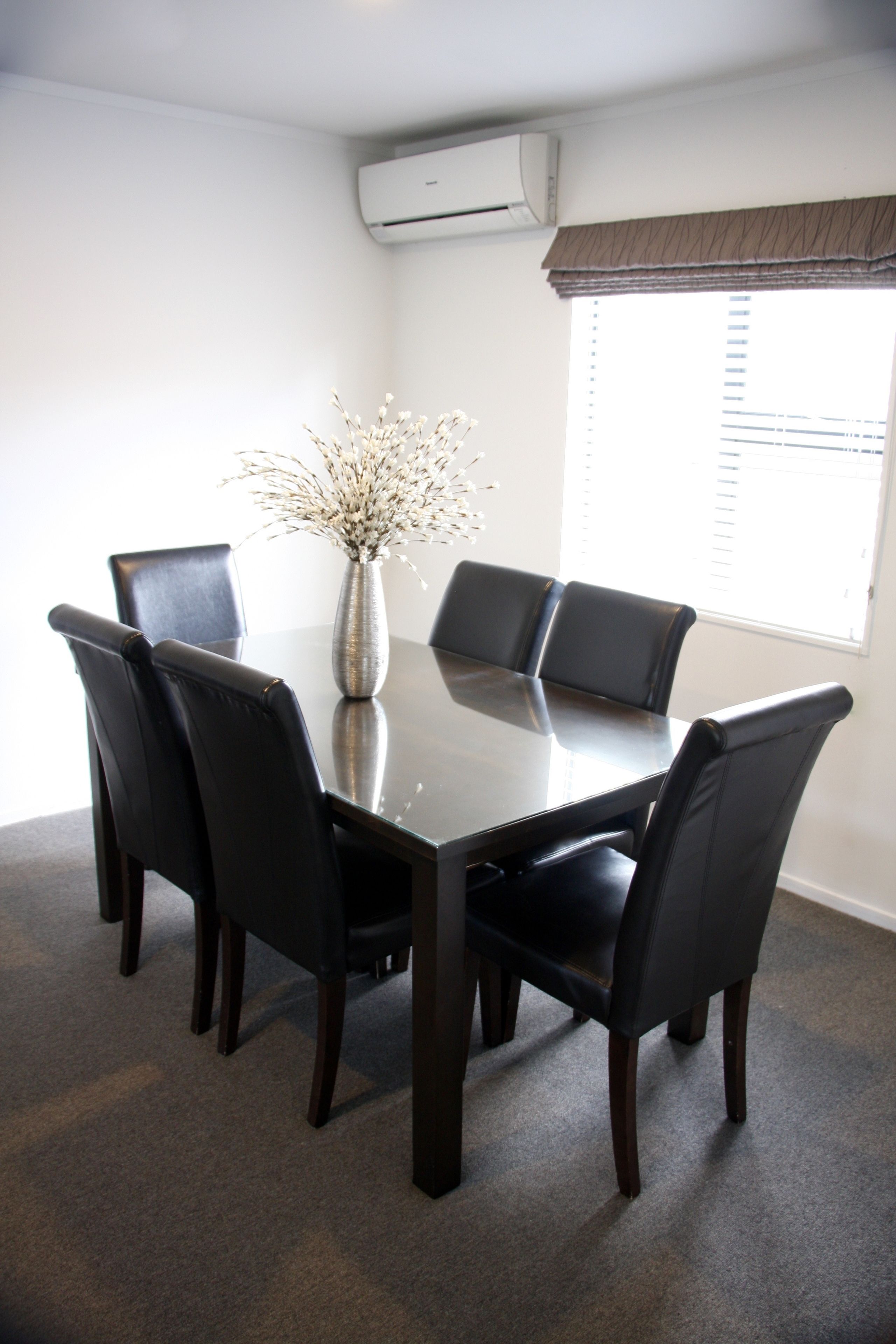 A conference room with a dark wooden table, six black leather chairs, a vase with white flowers, and an air conditioning unit on the wall.
