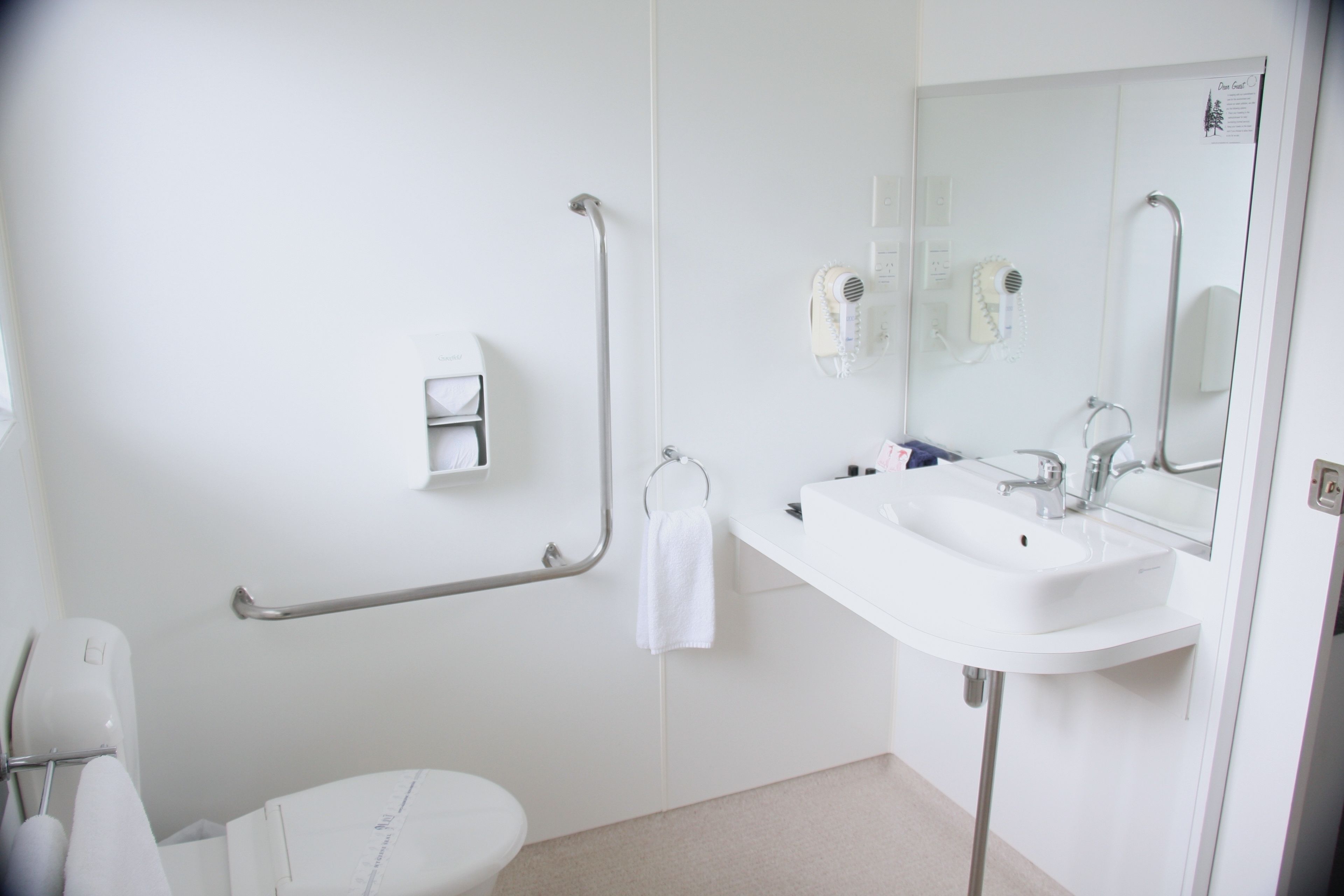 A modern bathroom with a white sink, mirror, and toilet. There is a grab bar next to the toilet and a towel rack above it.