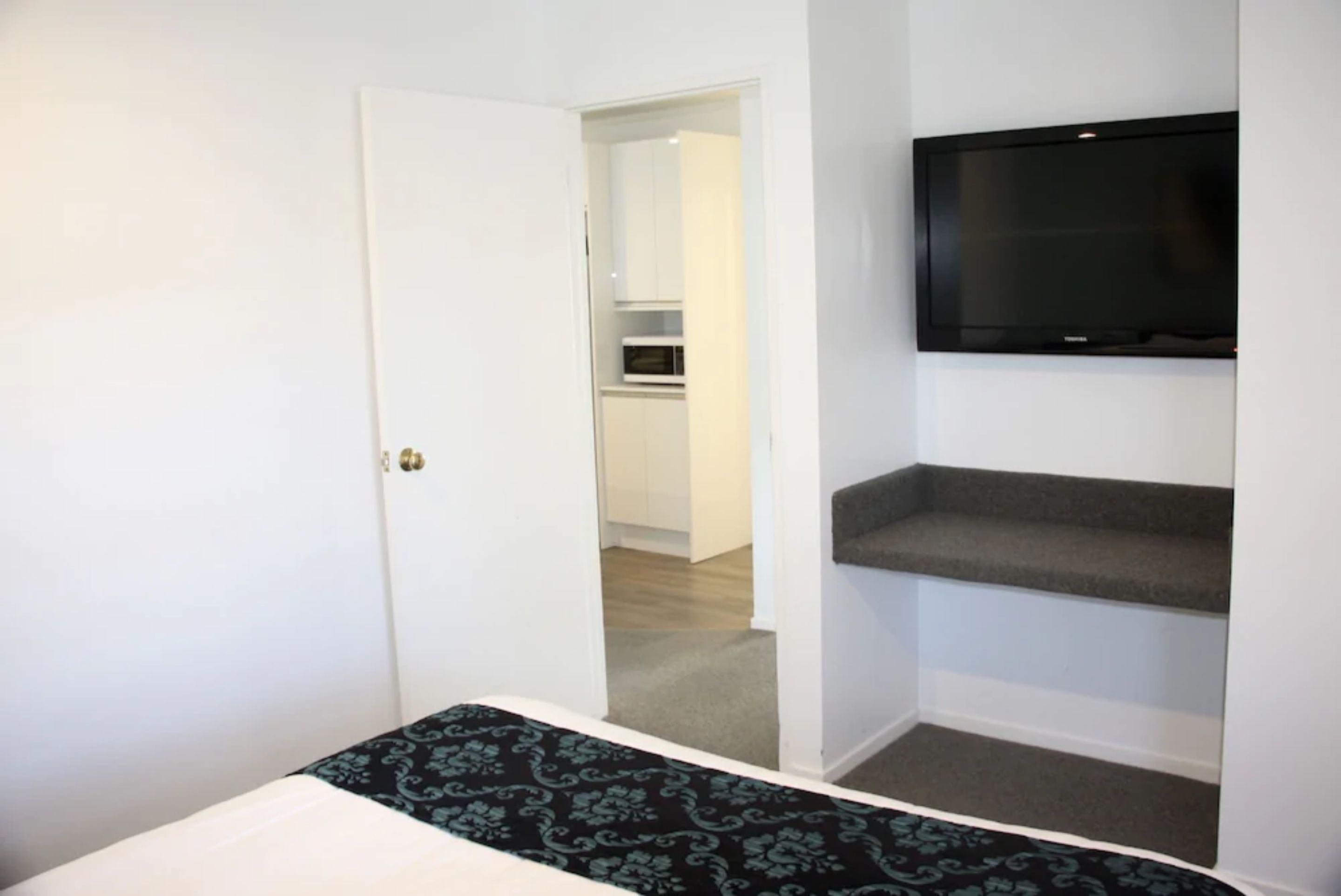 A bedroom with a bed, a TV mounted on the wall, and a door leading to a kitchen area.