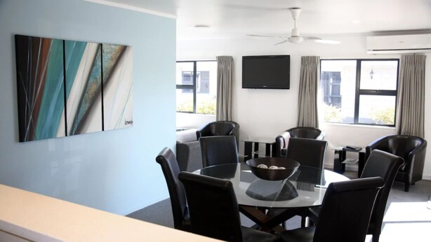 A modern dining area with a round table, black chairs, and a bowl of fruit. There is a ceiling fan, a flat-screen TV, and large windows with curtains. - Camelot on Ulster (Hamilton)