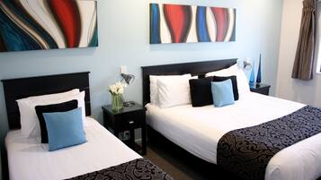 Habitación de hotel con dos camas, cada una con cabecero negro, ropa de cama blanca y almohadas azules. En las paredes, cuadros abstractos, una mesita de noche con un florero y una ventana con cortinas.