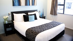 A neatly made bed with a patterned bedspread, a bedside table with a vase of flowers, and a wall-mounted abstract painting. - Camelot on Ulster (Hamilton)