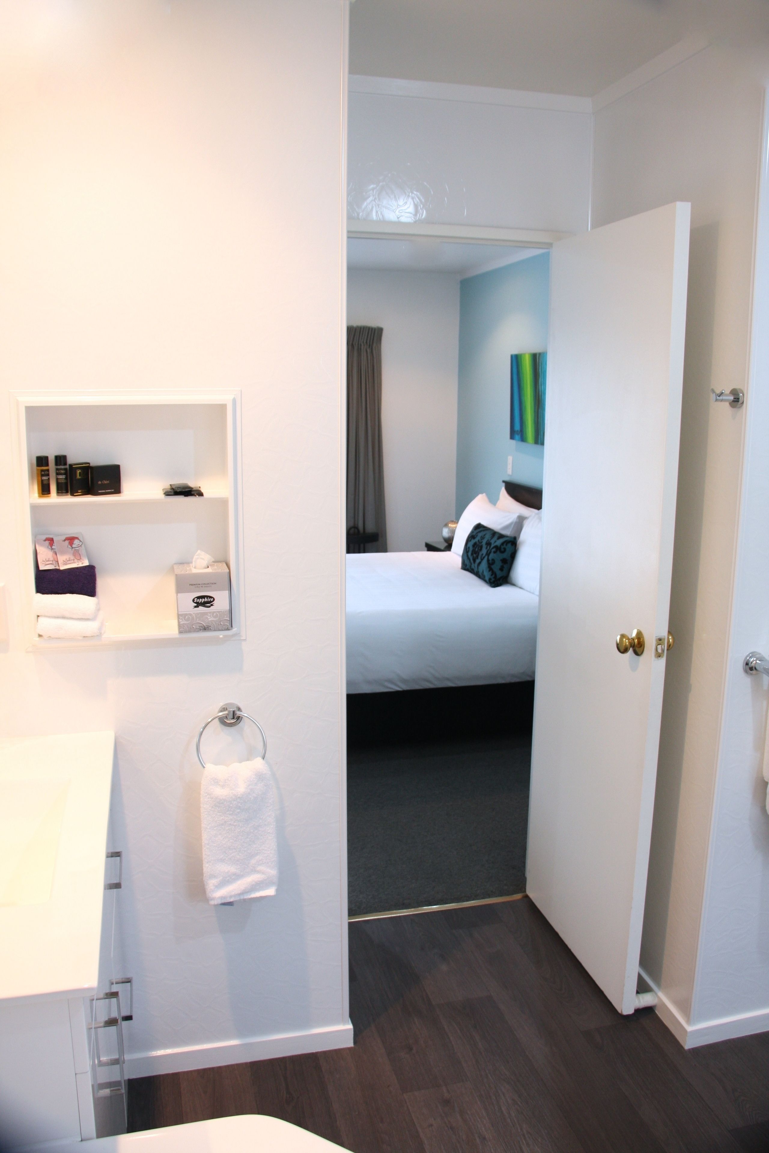 Quarto de hotel com cama, mesa de cabeceira, espelho, porta-toalhas e um pequeno armário com amenities.