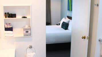 Quarto de hotel com cama, mesa de cabeceira, espelho, porta-toalhas e um pequeno armário com amenities.