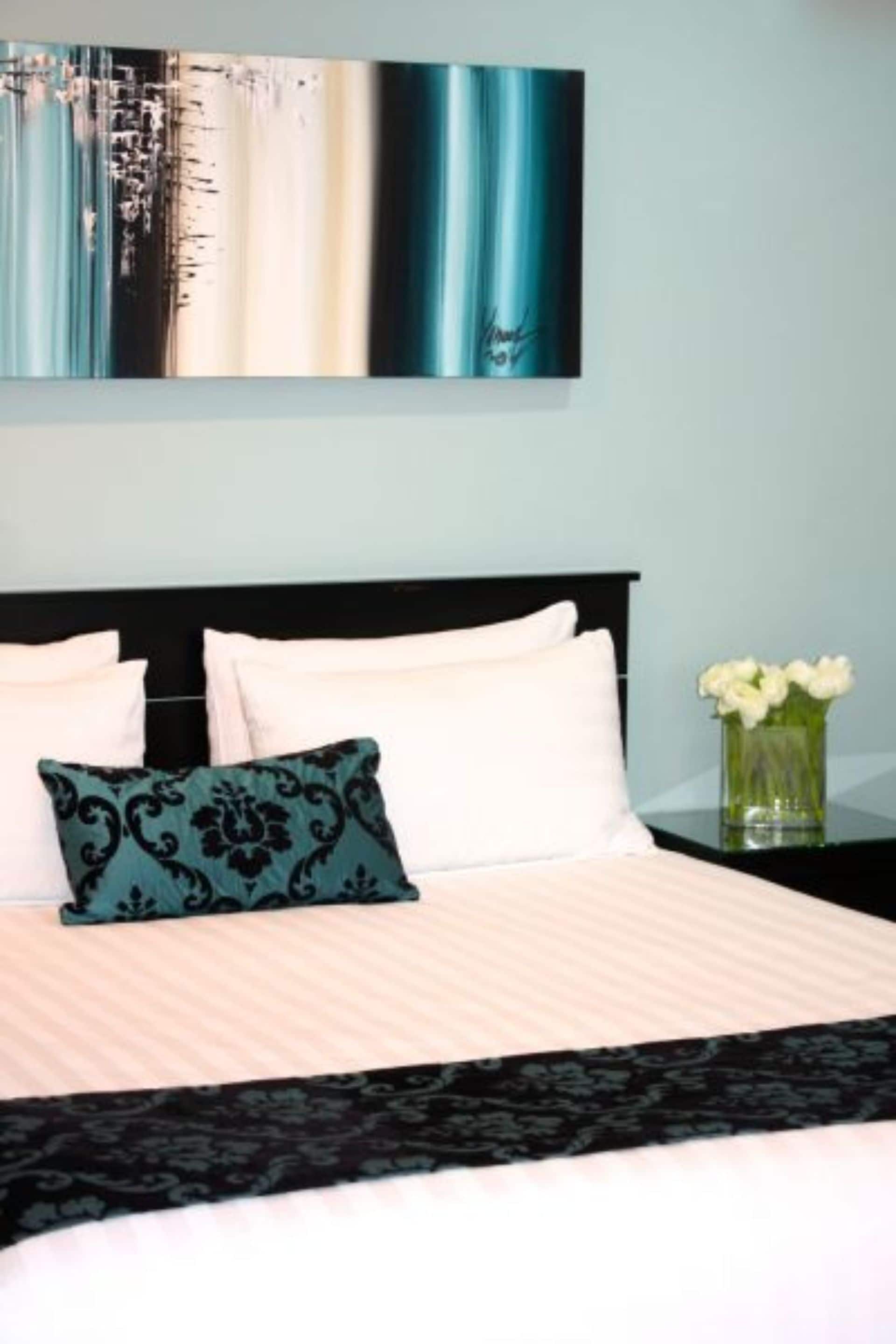 Un lit bien fait, avec des oreillers blancs et turquoise, une tête de lit noire et un vase rempli de fleurs blanches posé sur une table de chevet.