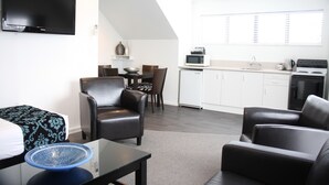 A modern living room with a flat-screen TV, a dining area with a table and chairs, a kitchen with white cabinets and appliances, and a large window with blinds. - Camelot on Ulster (Hamilton)