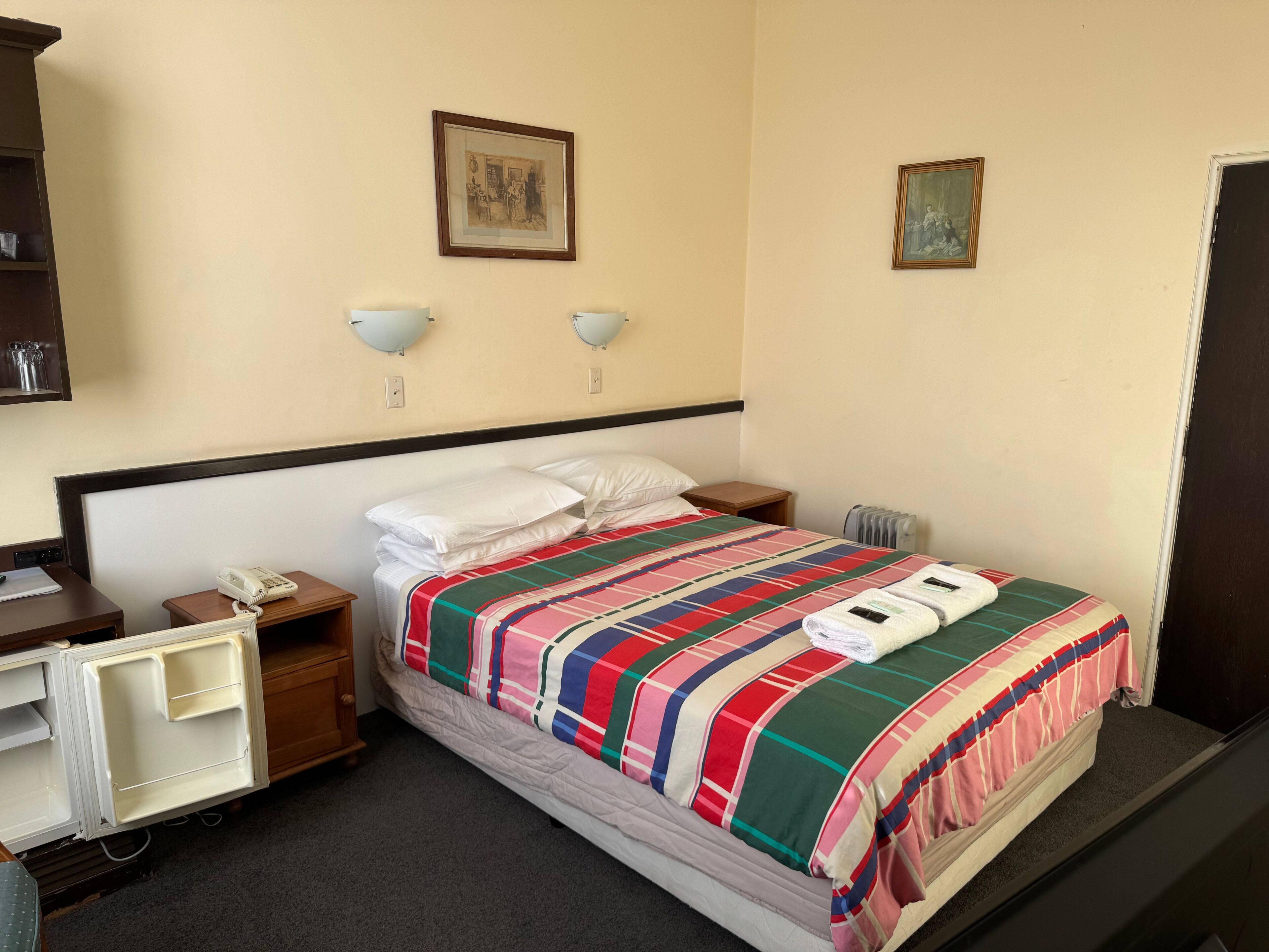 A hotel room with a bed, a bedside table with a phone, a small desk with a chair, and a shelf with items.