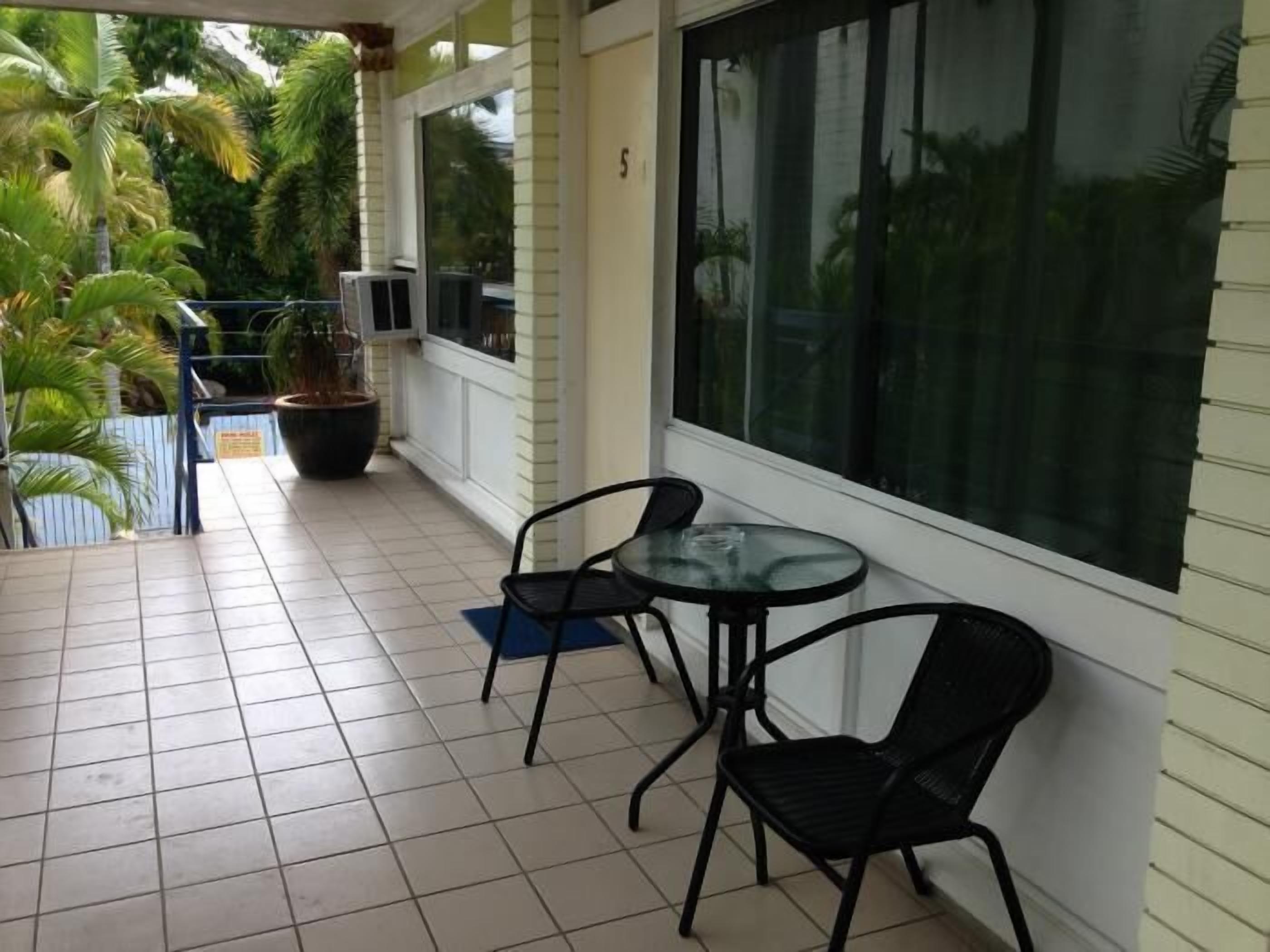 A covered patio area with tiled flooring, two black metal chairs, a glass table, and a potted plant.