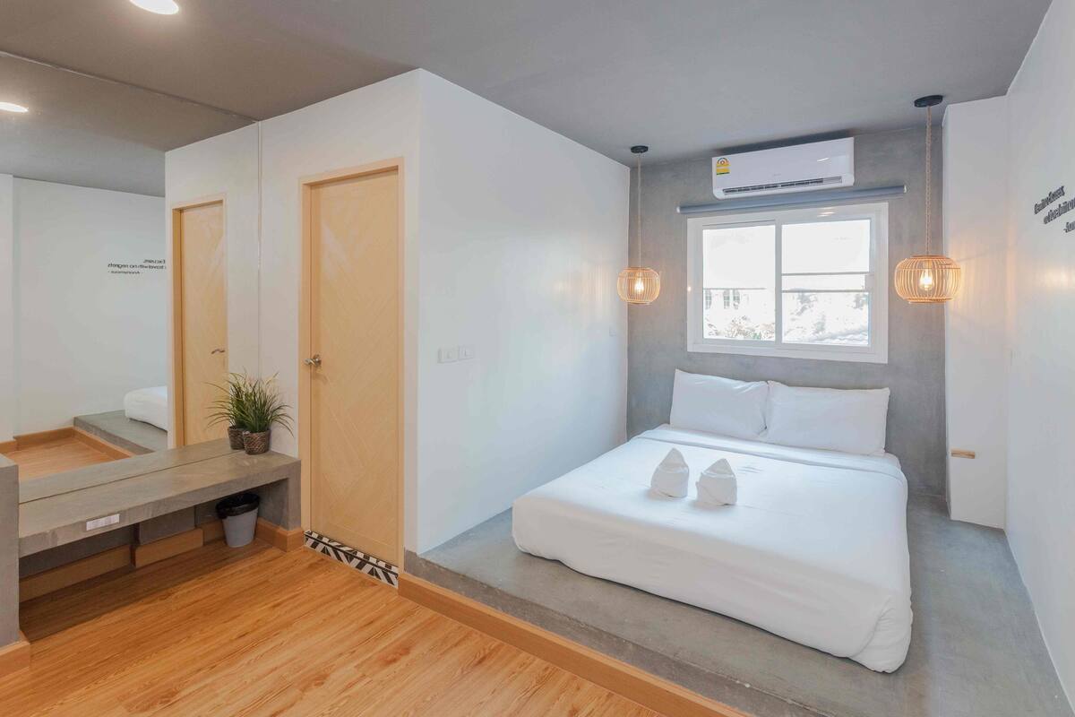 A modern hotel room with a bed, a small desk, a window, and a wall-mounted air conditioner.