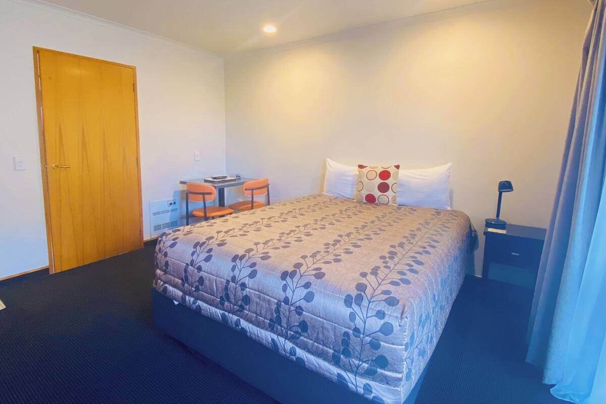 A hotel room with a bed, a desk with two chairs, a nightstand, and a door.