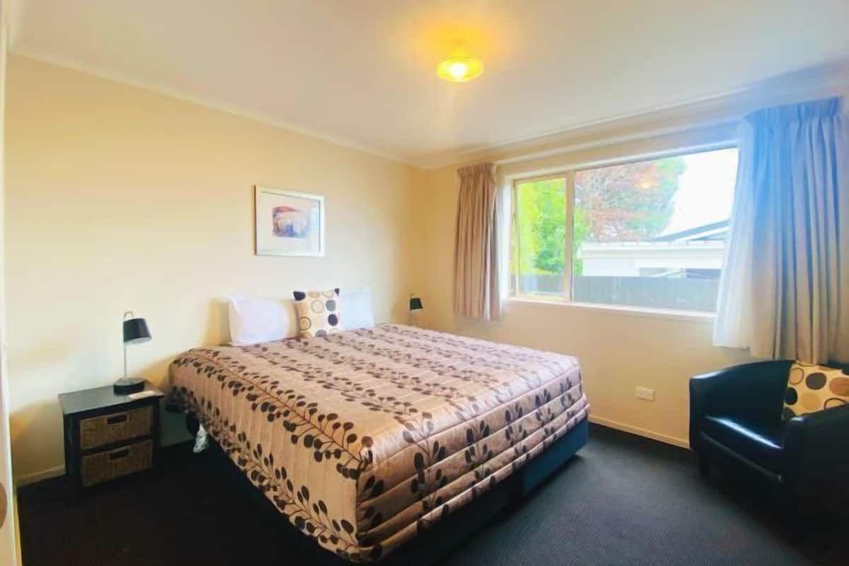 A hotel room with a bed, a nightstand, a chair, a window with curtains, and a picture on the wall.