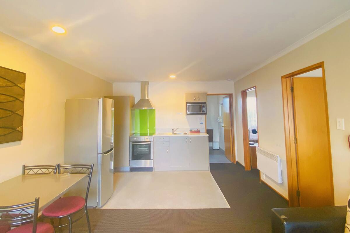 A compact kitchen with a refrigerator, oven, microwave, and a green range hood. There is a dining area with a table and chairs, and a small kitchenette in the background.
