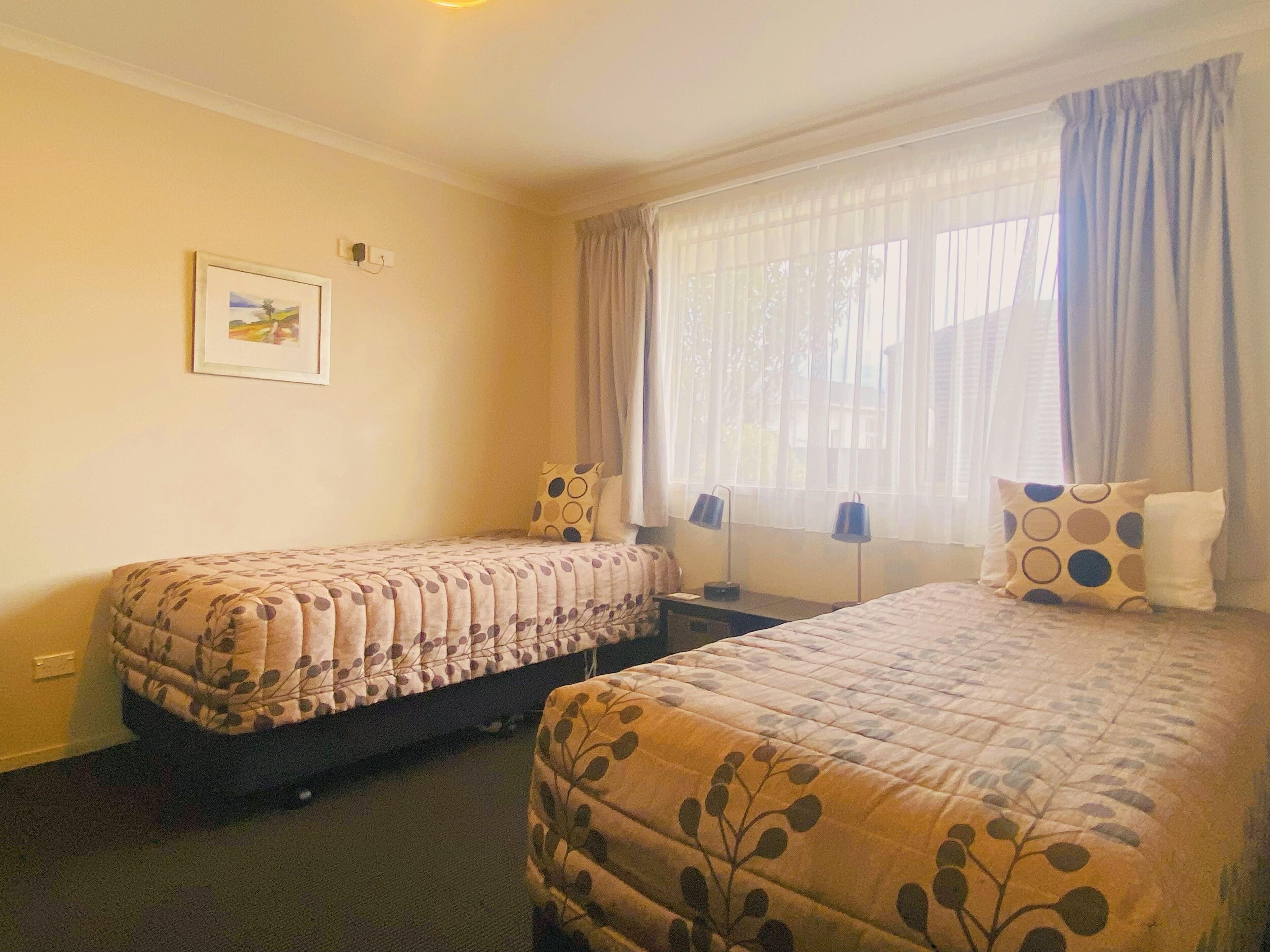 Una habitación de hotel con dos camas, una ventana con cortinas, un cuadro en la pared y una mesita de noche con lámparas.