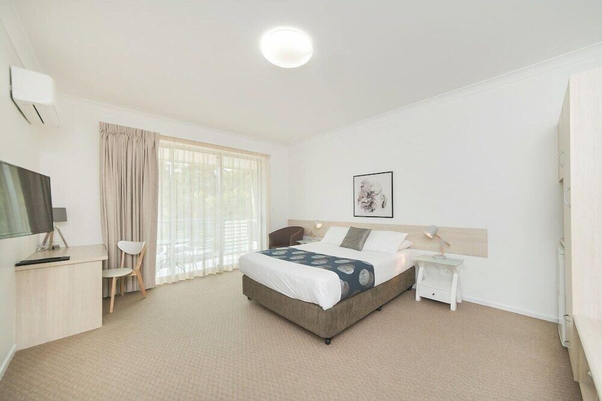 A hotel room with a bed, a desk, a chair, a TV, a bedside table, and a window with curtains.