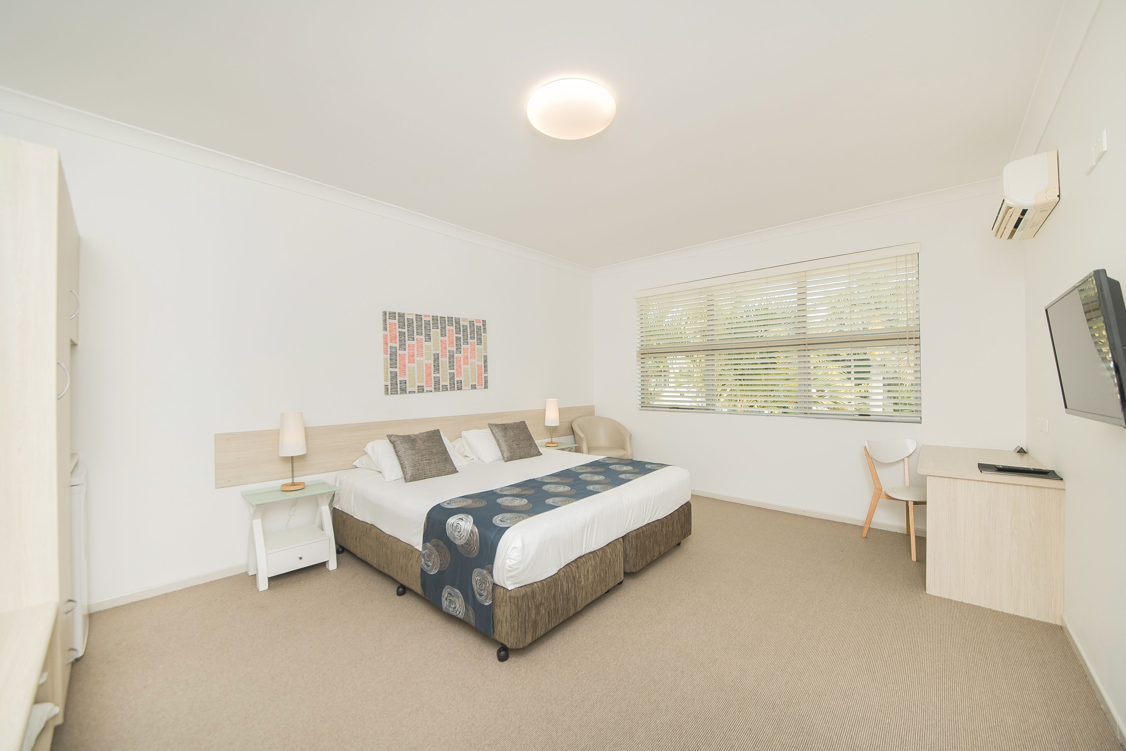 A bedroom with a bed, a desk, a chair, a nightstand, a lamp, a window with blinds, and a ceiling light.