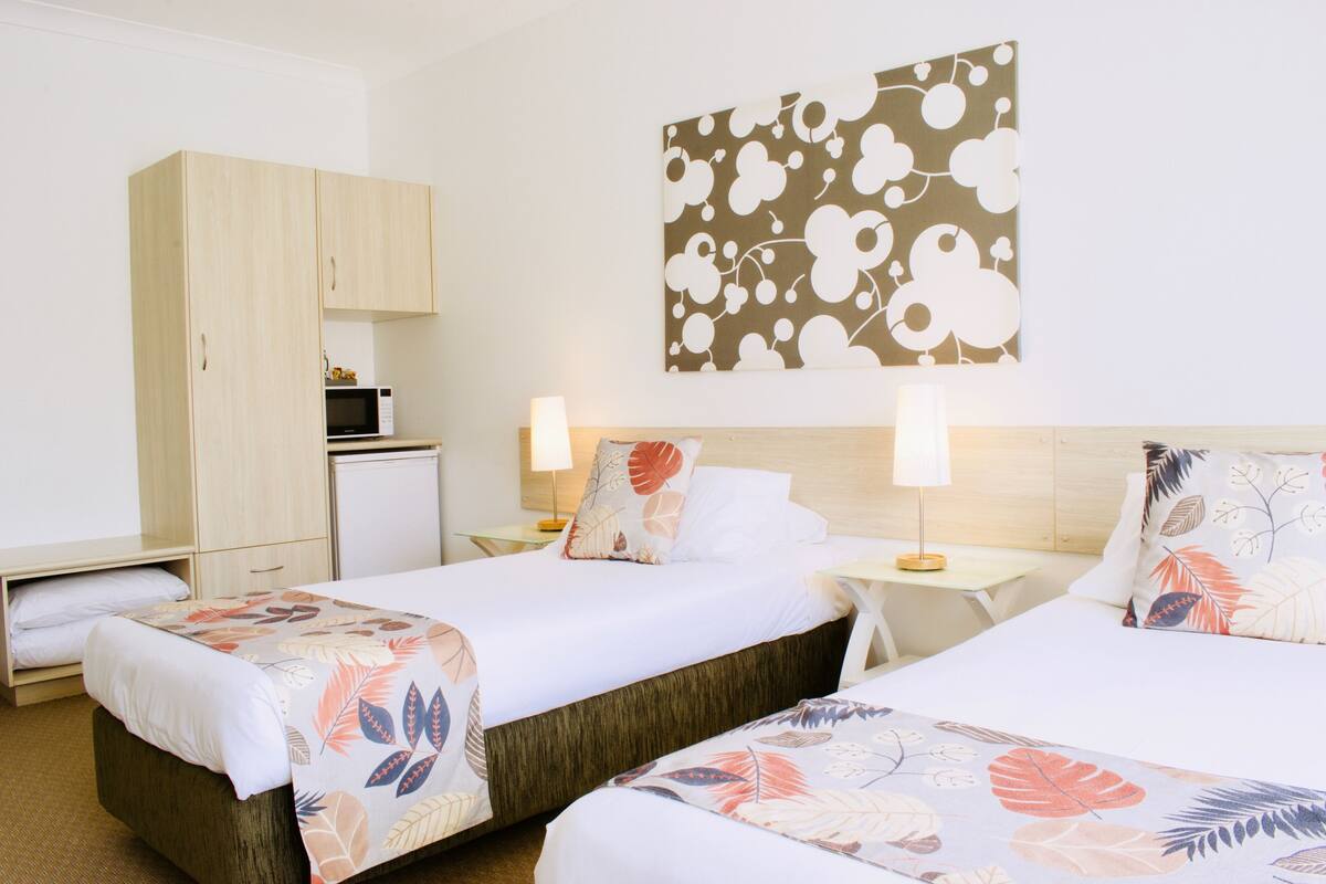 A hotel room with two beds, a wooden wardrobe, a microwave, and a floral artwork on the wall.