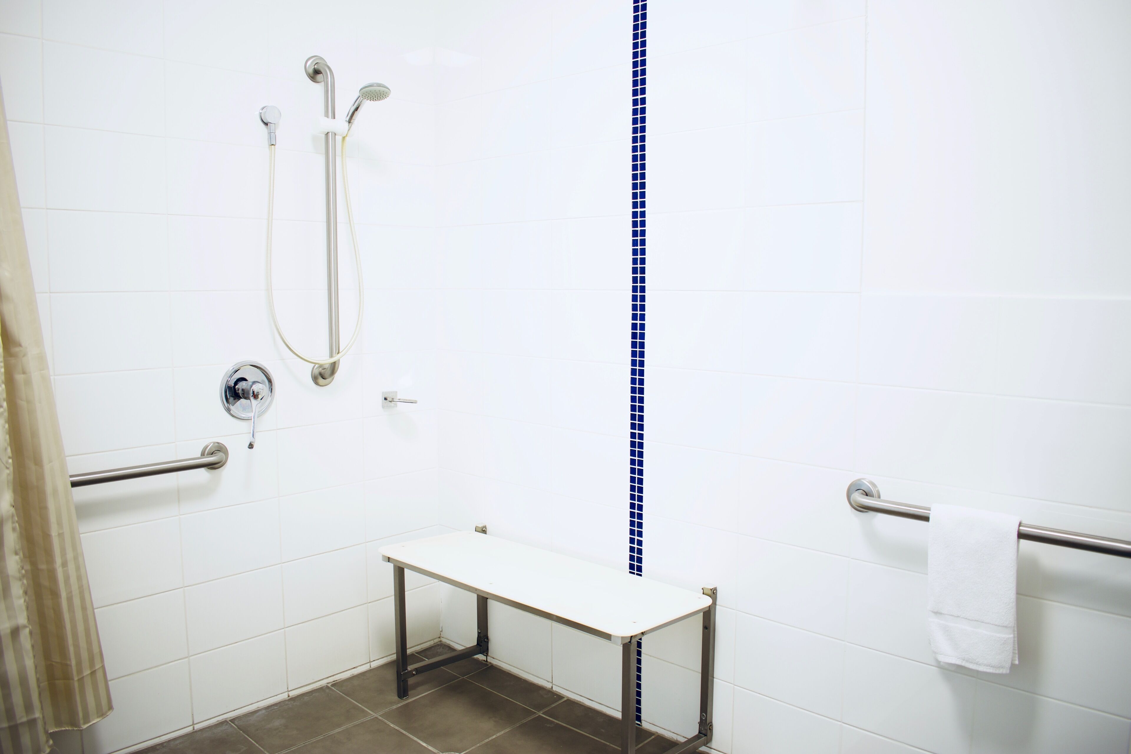 Un bagno con pareti rivestite di piastrelle bianche, una fascia verticale di piastrelle blu, una panca bianca, un portasciugamani e una doccia con soffione manuale.