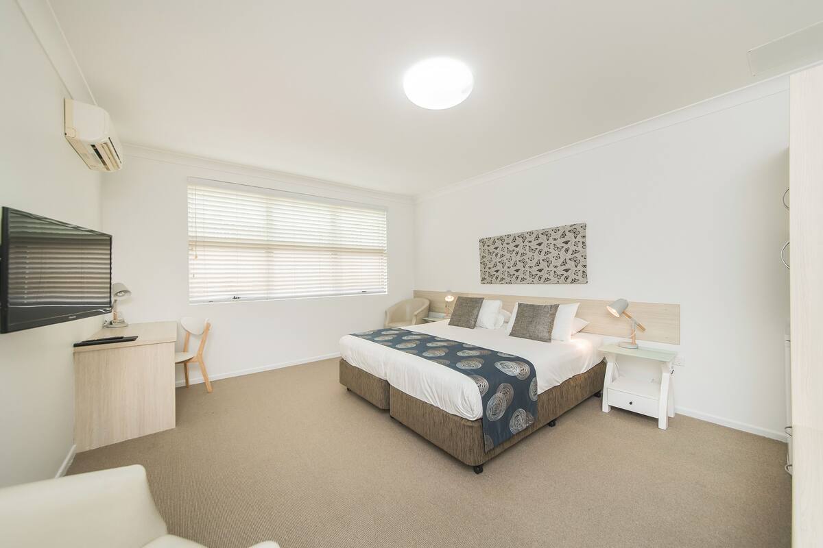 A bedroom with a bed, a desk, a chair, a TV, and a window with blinds.