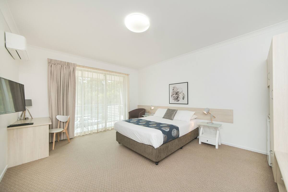 A hotel room with a bed, a desk, a chair, a TV, a bedside table, and a window with curtains.