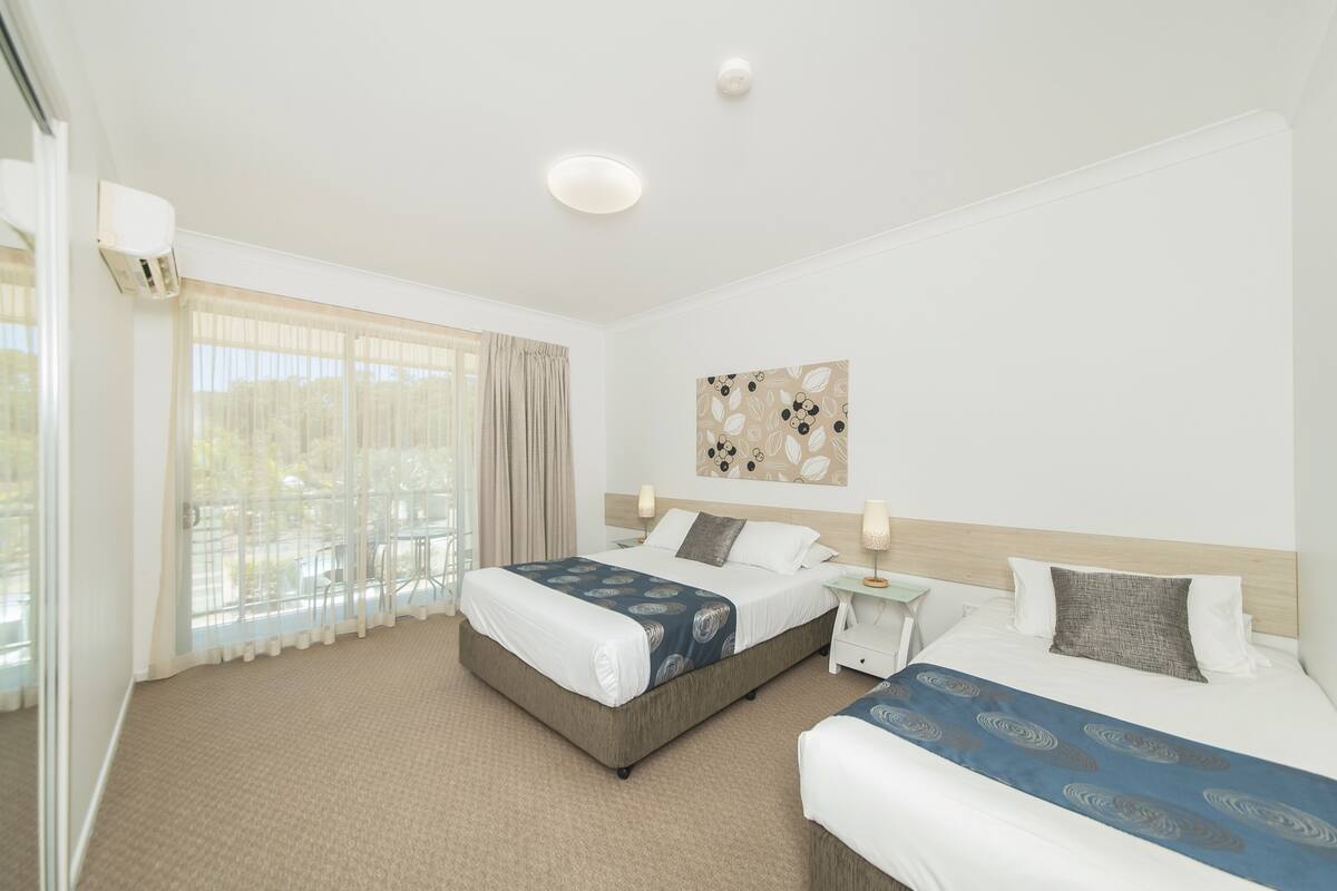 A hotel room with two beds, a large window with curtains, a ceiling light, and a wall-mounted artwork.
