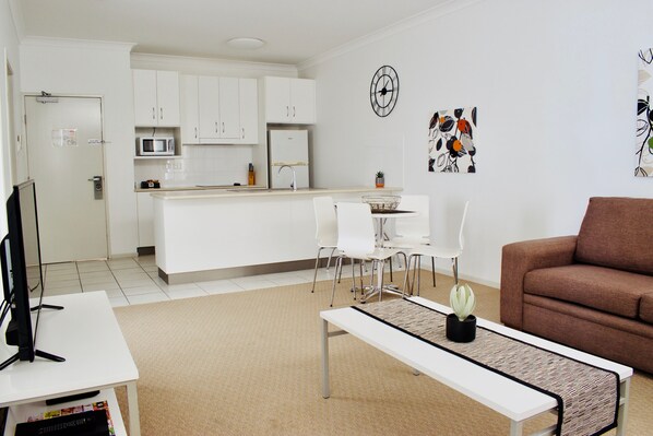 Un salon moderne avec une table basse blanche, un canapé marron et un téléviseur posé sur une table blanche. On aperçoit, au fond, une cuisine avec des meubles blancs et un four à micro-ondes.