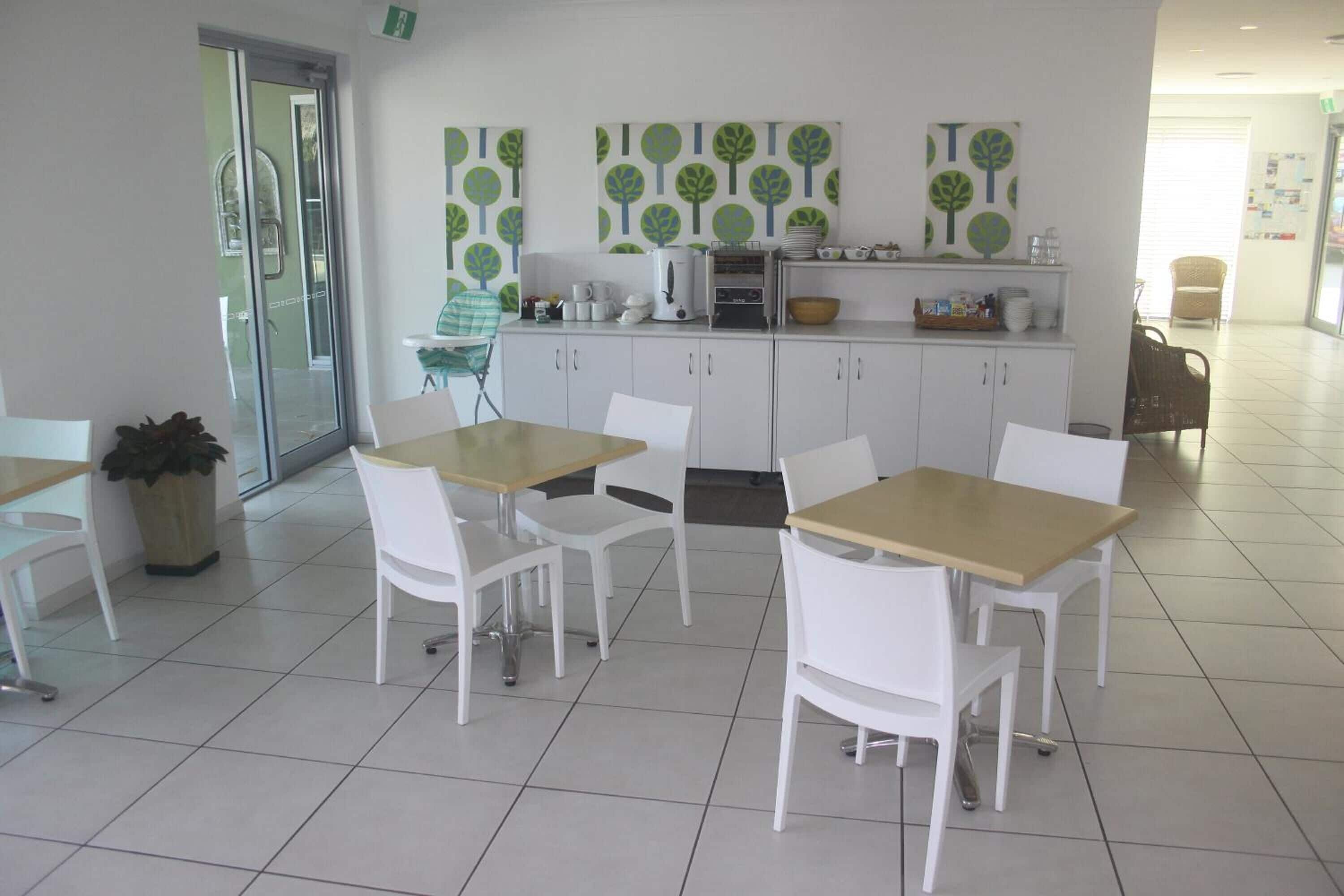 Un comedor limpio y moderno con sillas y mesas blancas, una máquina de café y elementos decorativos en la pared.