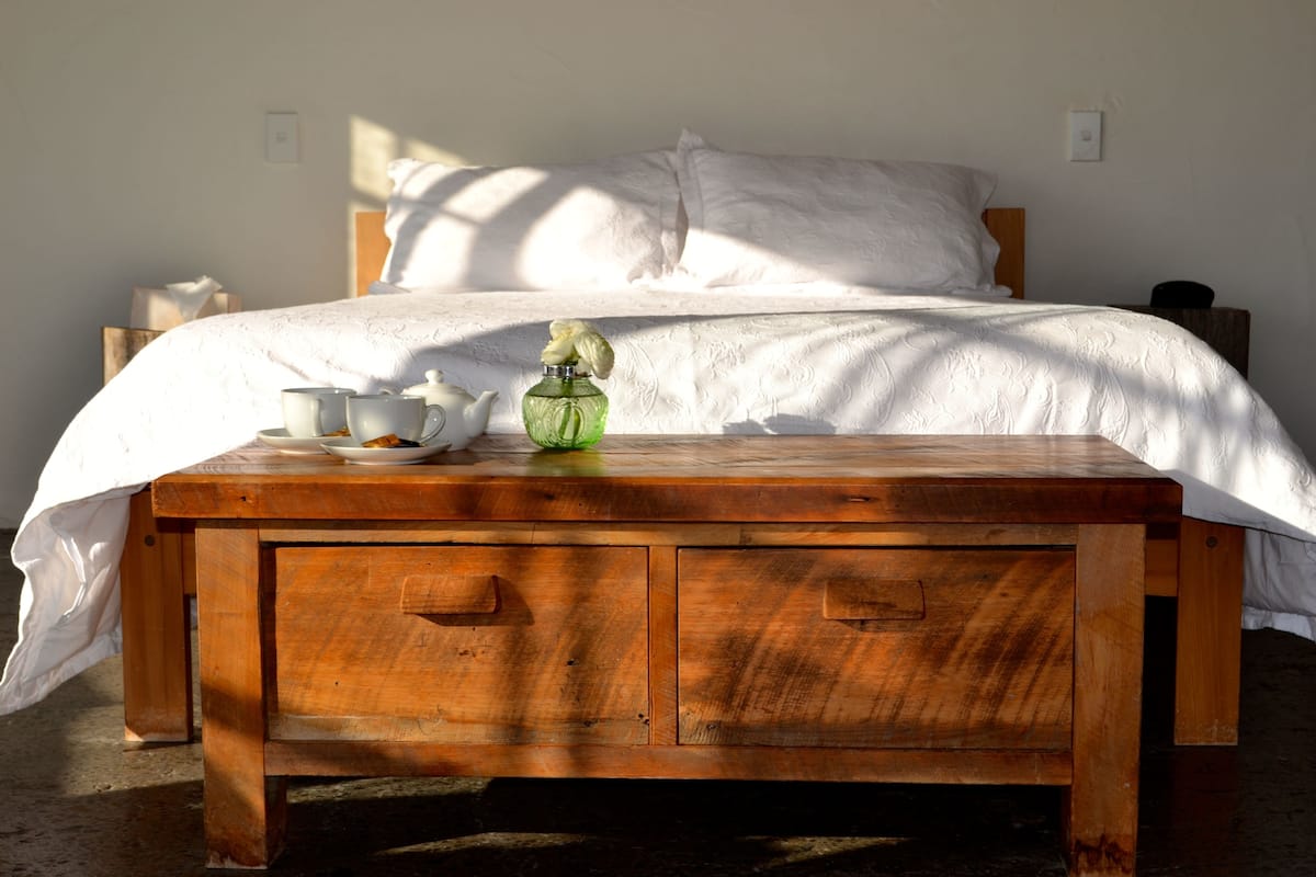 A neatly made bed with white linens, a wooden bedside table with a teapot, cups, and a green bottle.