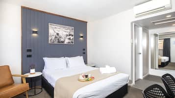 Una habitación de hotel moderna con una cama, una pequeña mesa redonda con un jarrón de flores, un sillón de cuero marrón y una silla negra, junto a un espejo grande.