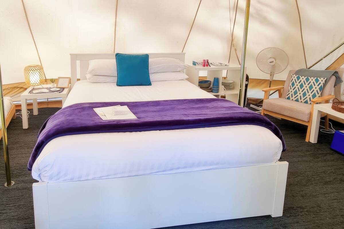 Een bed met een wit frame, een paars dekbed en een blauw kussen. Er staat een klein tafeltje met een lamp en een mandje.