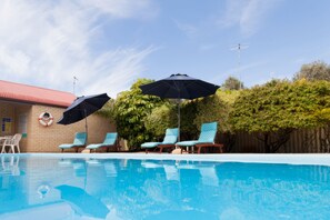 A swimming pool with lounge chairs and umbrellas, surrounded by greenery and a building with a red roof. - Busselton Villas & Glamping Village (West Busselton)