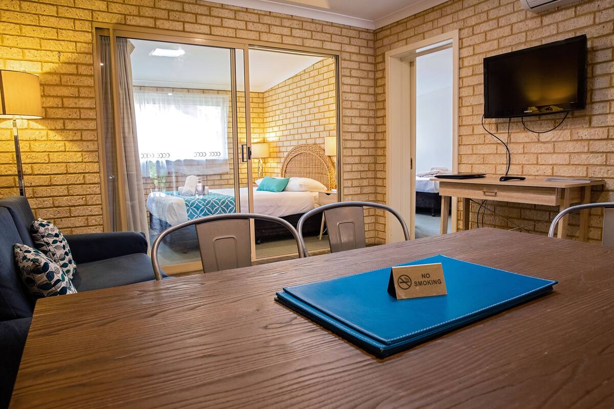 Een hotelkamer met een houten tafel, een blauwe map met een ‘Niet roken’-bordje, een televisie aan de muur en uitzicht op het balkon met een bed en een rieten stoel.