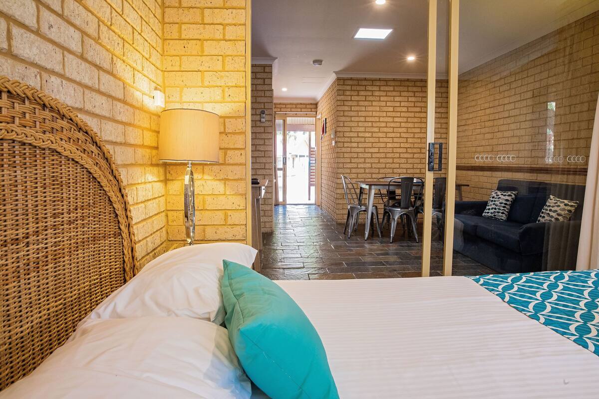 Hotelkamer met een bakstenen muur, een rieten hoofdbord, een bed met wit en teal beddengoed, een glazen scheiding, een zithoek met stoelen en een tafel, en een bank met gekleurde kussens.